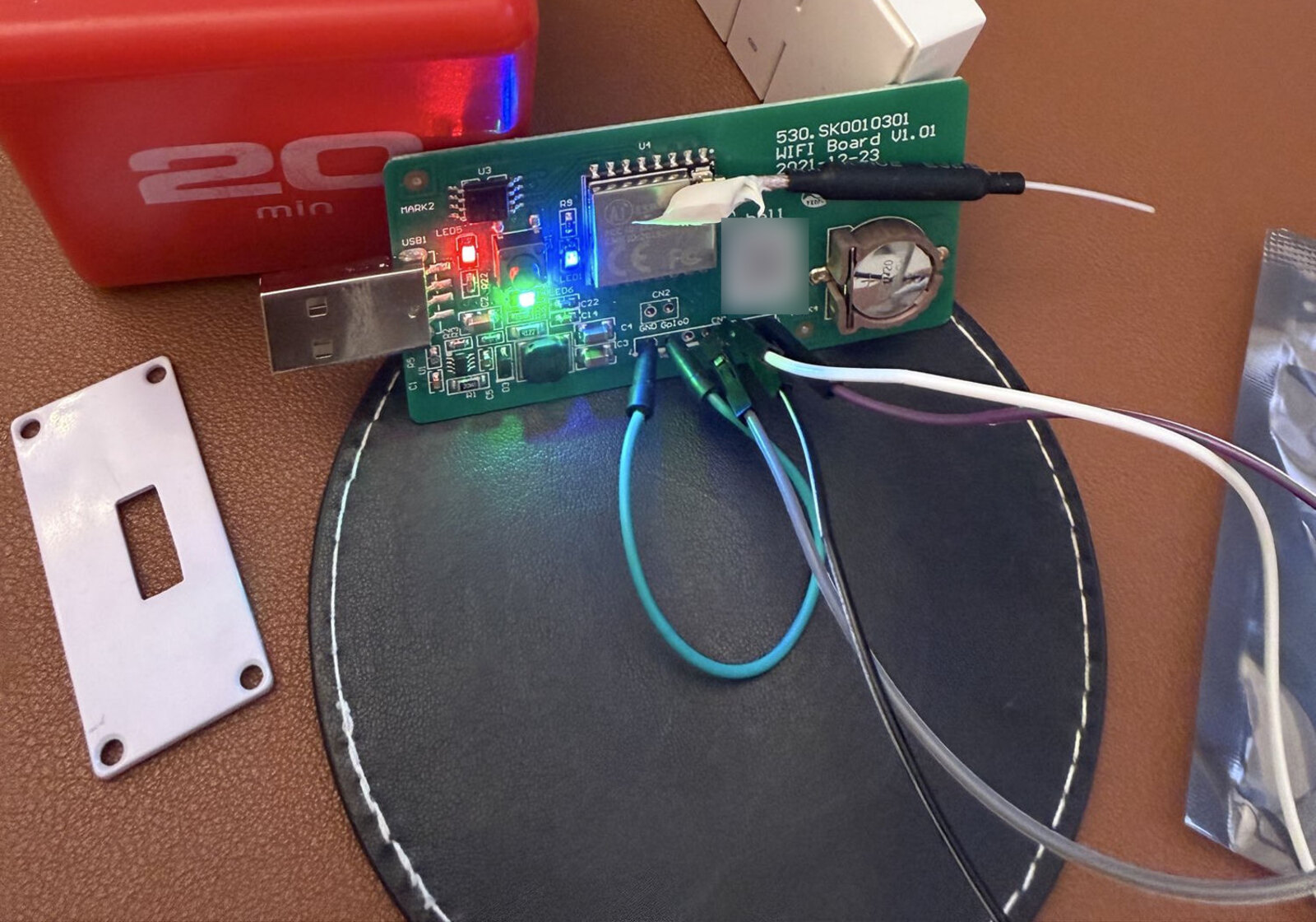 The old WiFi logger board out of its case on a desk, USB plug on the left, three status LEDs lit, four jumper leads clipped to the header on the right, with a red kitchen timer and a white remote behind it.